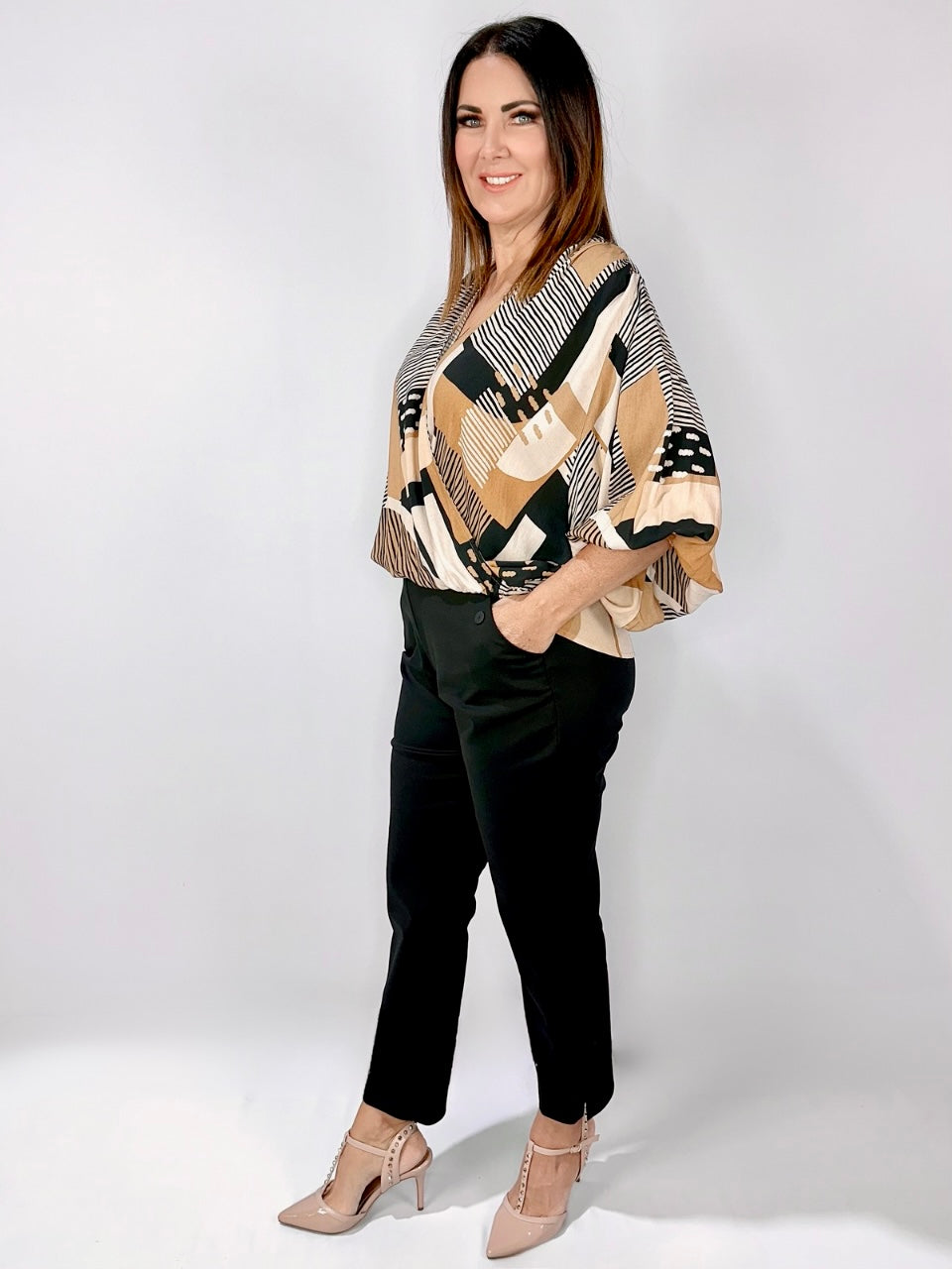 A woman stands confidently in a stylish printed blouse, black pants, and nude heels against a clean white backdrop.