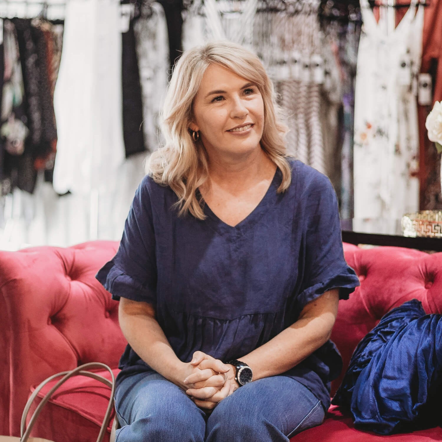 A woman in a navy top and jeans sits on a red sofa, surrounded by clothing racks, with a handbag beside her.