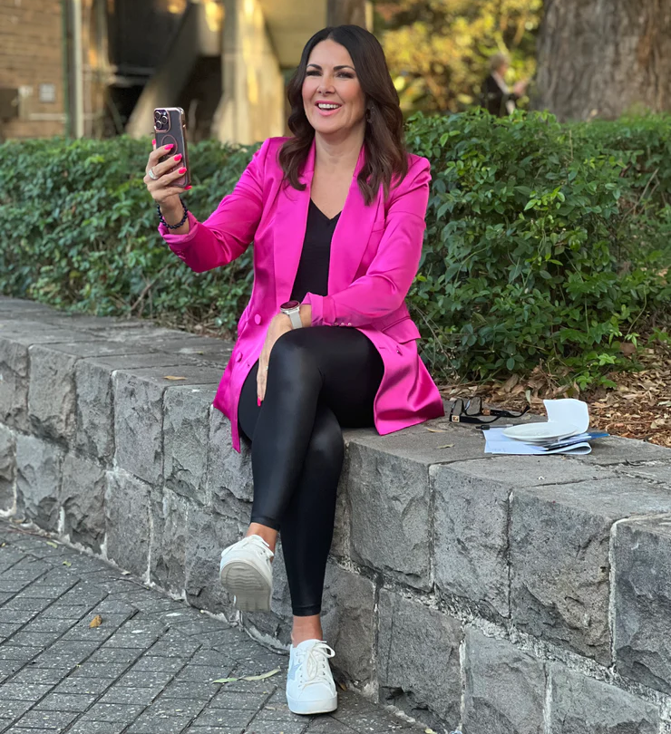 A woman in a vibrant pink blazer smiles while taking a selfie outdoors, seated casually on a stone ledge.