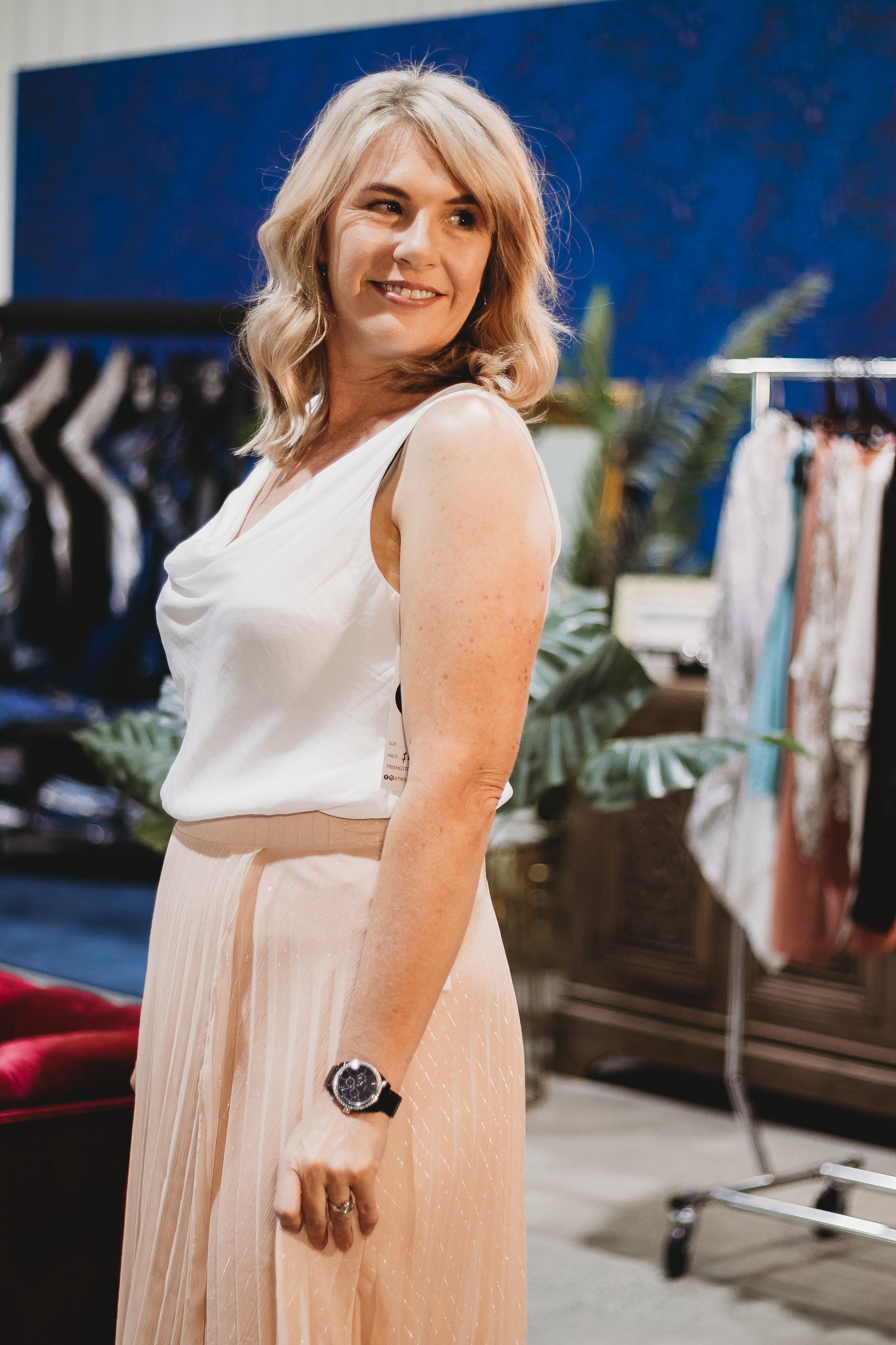 A woman confidently showcases her outfit during a styling session, with clothing racks and a boutique setting in the background.