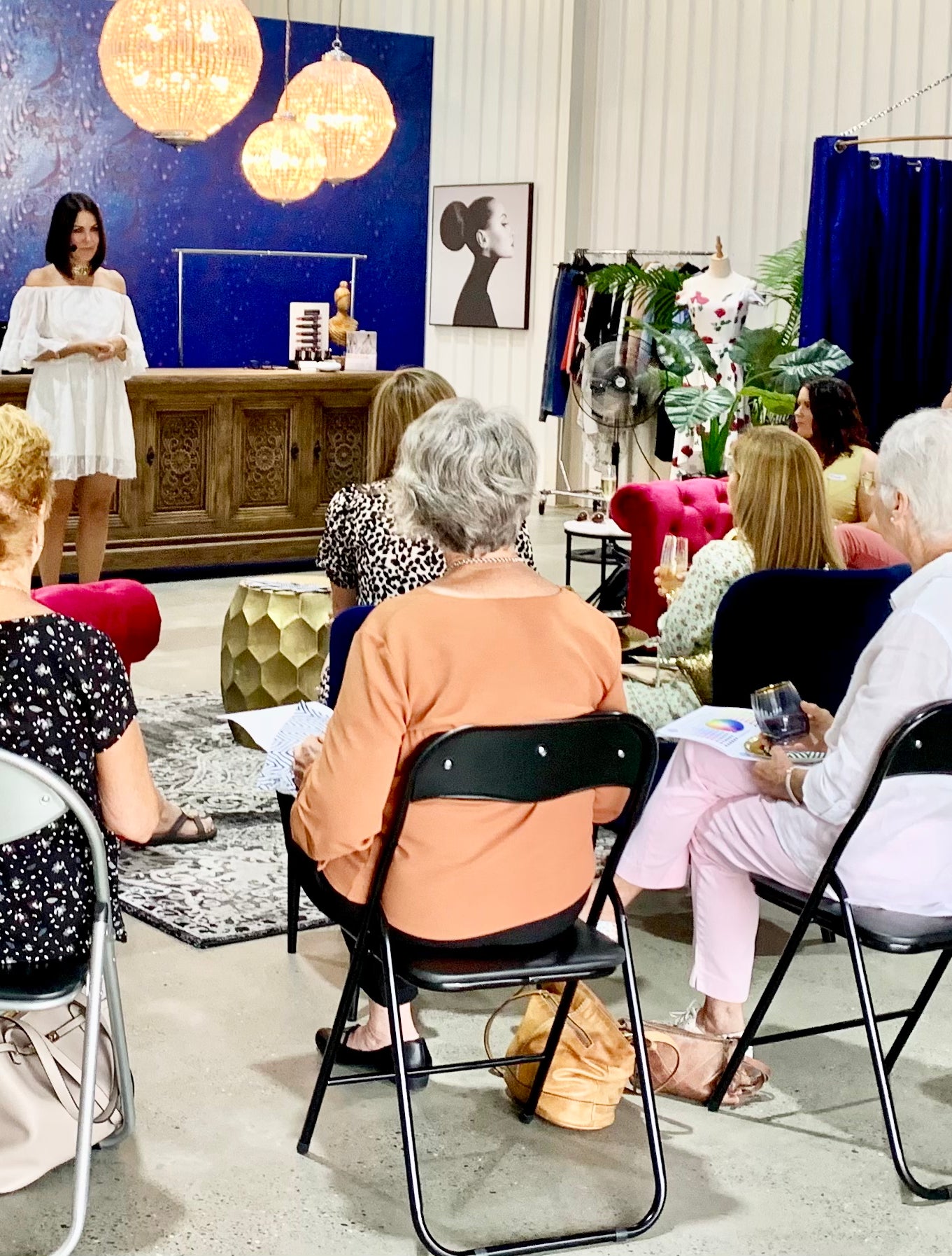 A group of women attends a styling session, seated in rows, while the presenter shares fashion tips in an elegant, vibrant studio space.