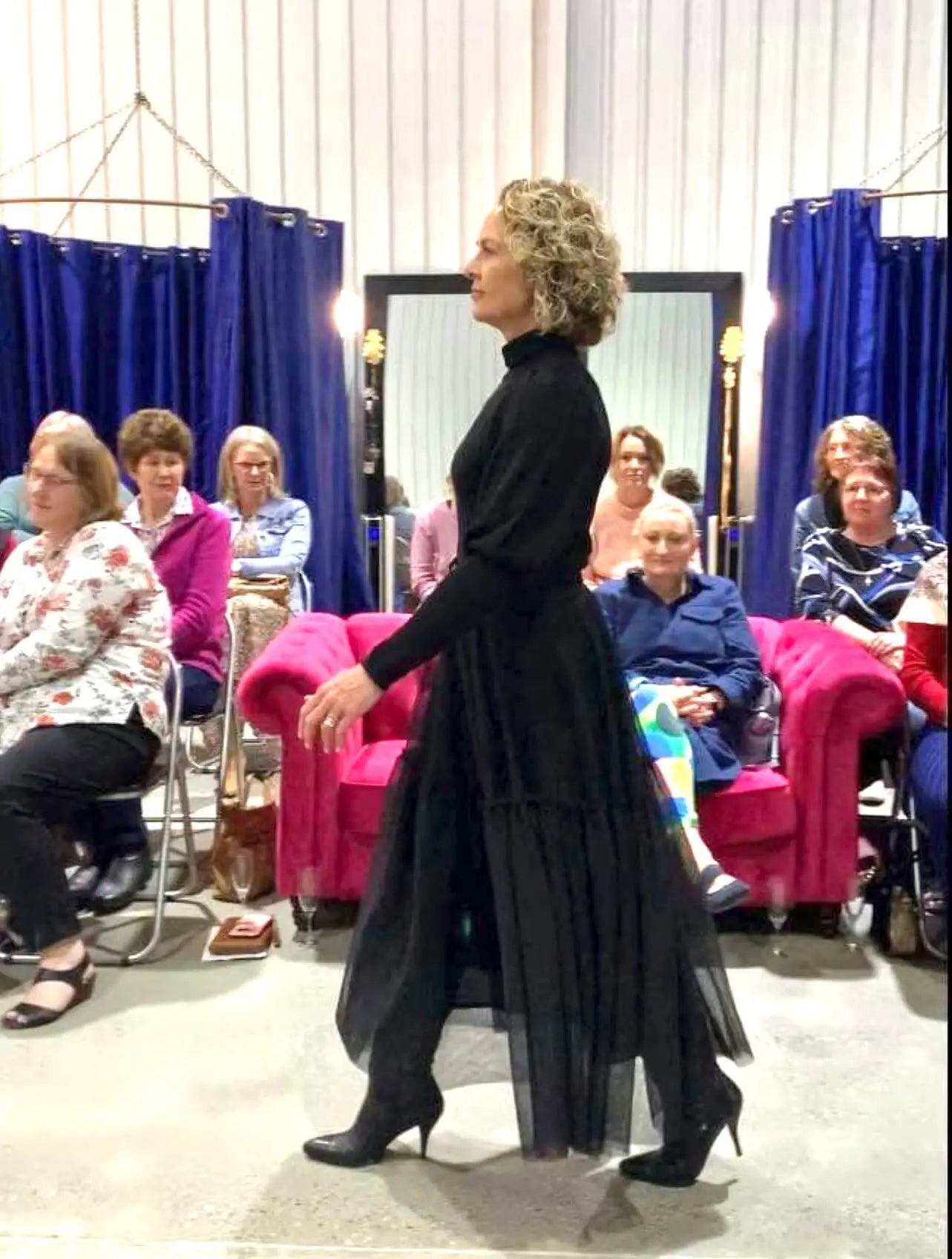 A model walks confidently in a black outfit during a style presentation, with an audience seated in the background.