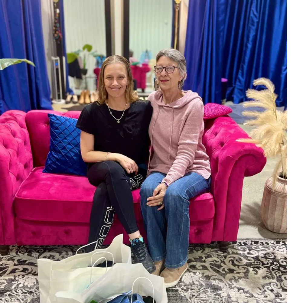 Two smiling women sit on a bright pink couch surrounded by shopping bags, in a stylish studio setting.