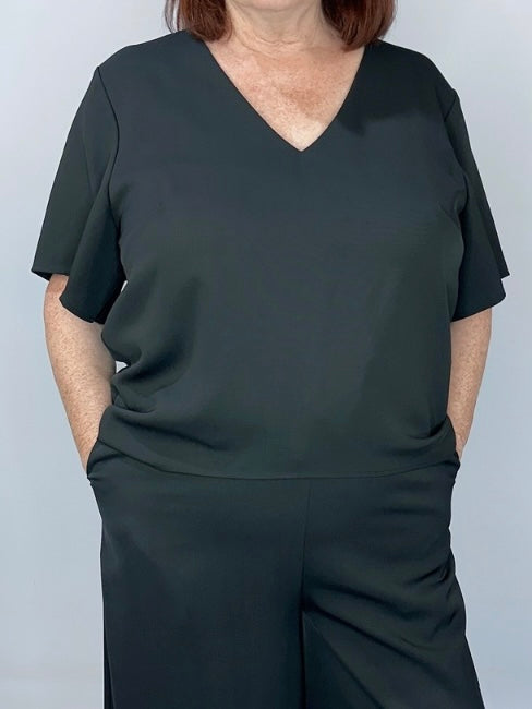 Woman wearing a black short-sleeve V-neck top and black wide-leg pants, standing with hands in pockets.