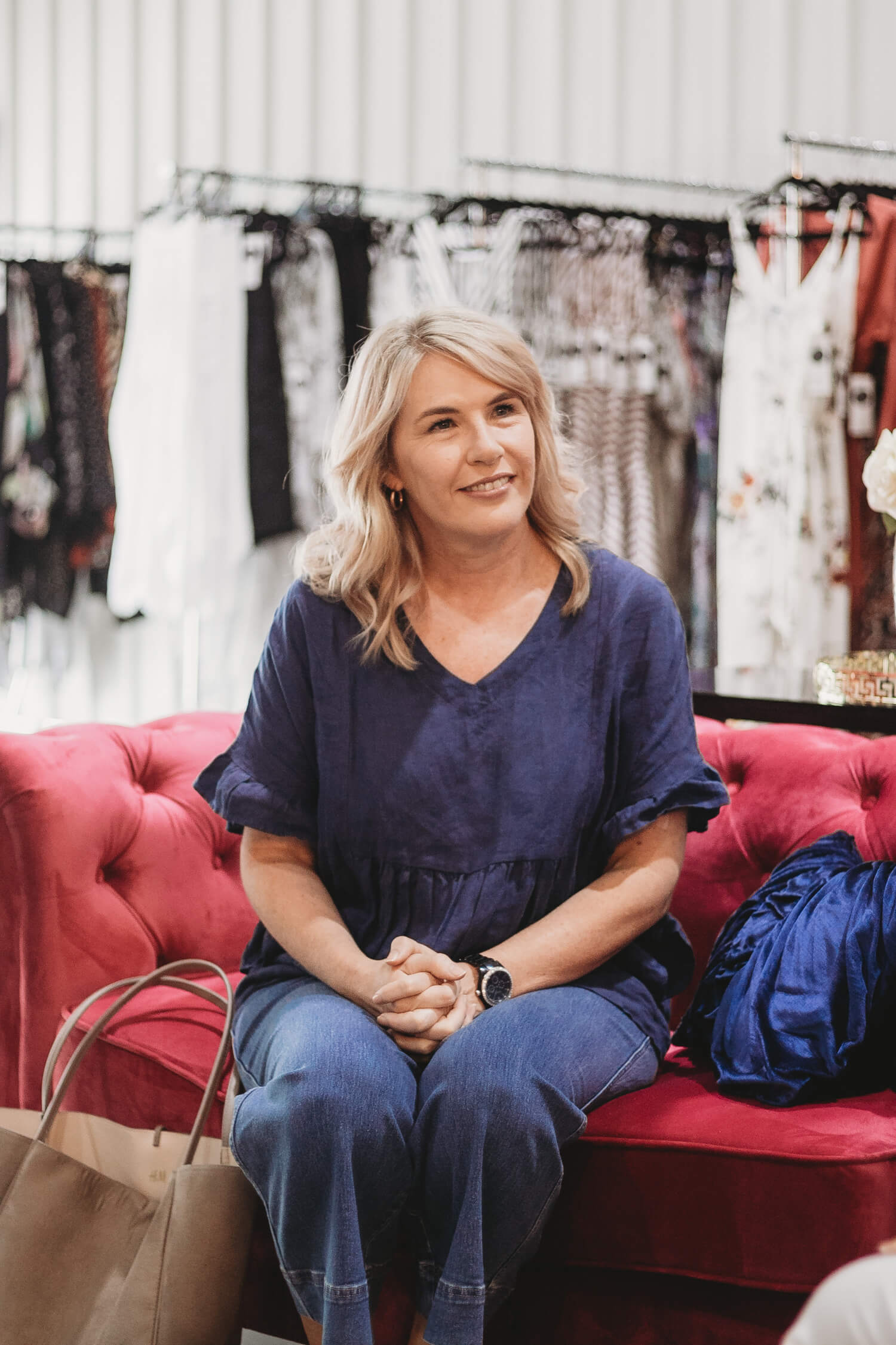 A woman in a navy top and jeans sits on a red sofa, surrounded by clothing racks, with a handbag beside her.