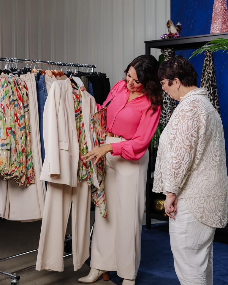 A stylist in a bright pink blouse helps a client select an outfit from a rack of neutral and colourful pieces.
