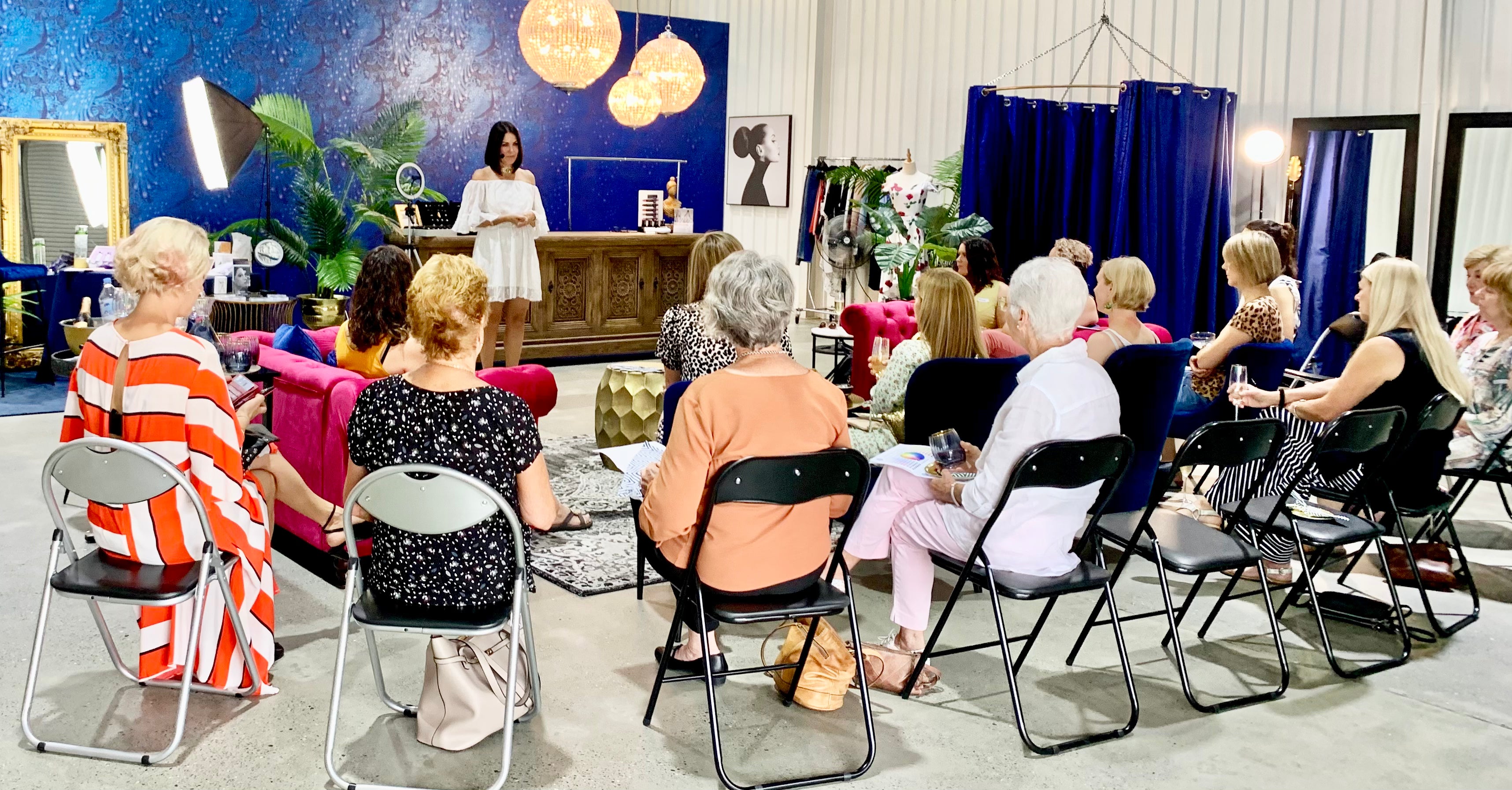 A group of women attends a styling session, seated in rows, while the presenter shares fashion tips in an elegant, vibrant studio space.