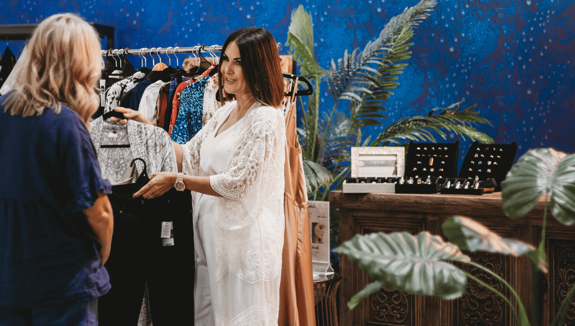 A stylist assists a client with outfit selection in a boutique, surrounded by racks of clothing and elegant decor.