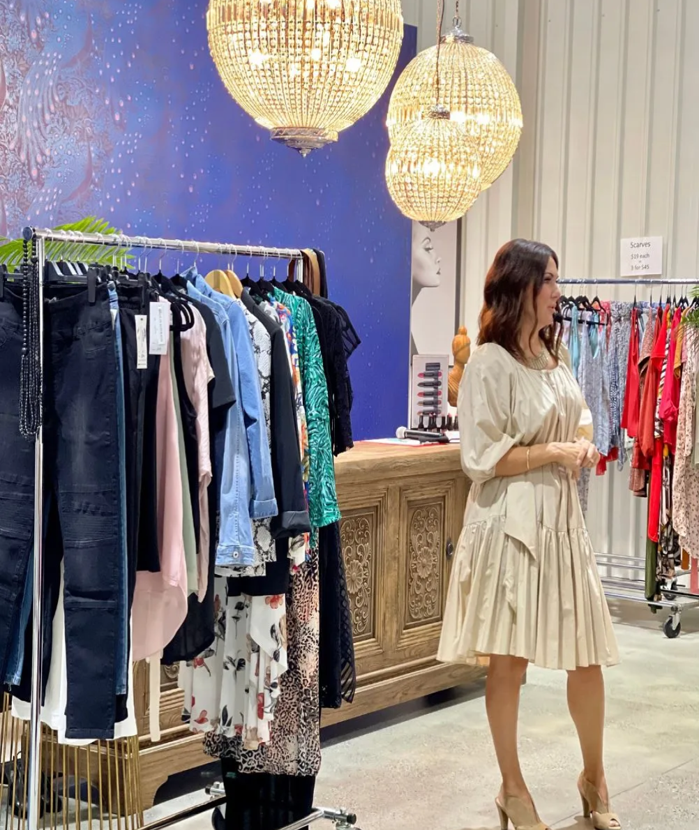 A stylist showcases a neutral-toned dress surrounded by racks of vibrant clothing under stunning chandeliers.