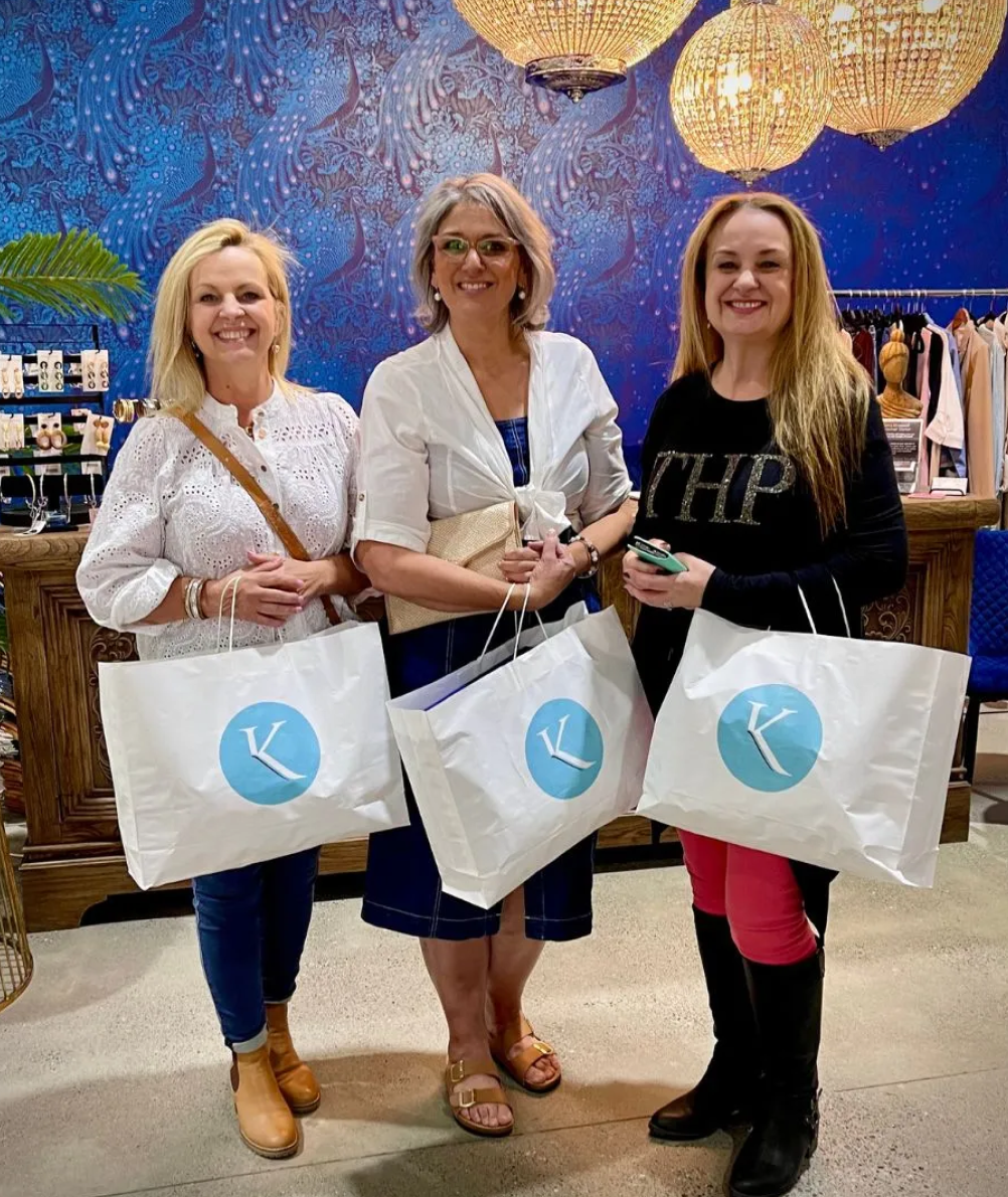 Three women holding shopping bags, smiling in front of a vibrant blue background.