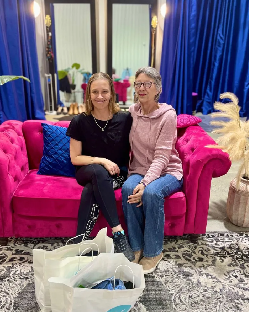 Two smiling women sit on a bright pink couch surrounded by shopping bags, in a stylish studio setting.